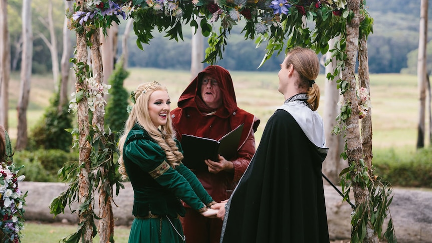 Man dressed in maroon, hooded velvet cloak standing behind couple dressed in medieval clothes standing under a flowery altar.