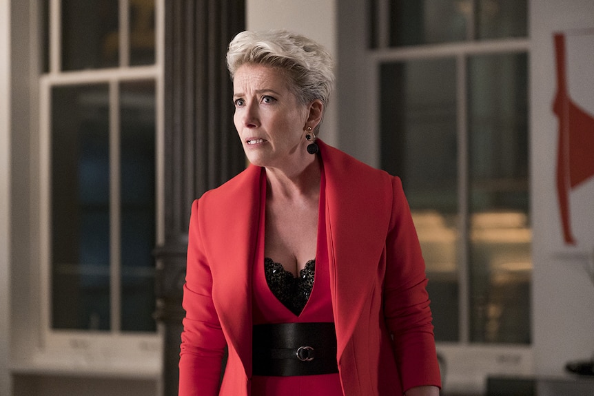 Emma Thompson, with a concerned expression, stands in an apartment, wearing a red pant suit.
