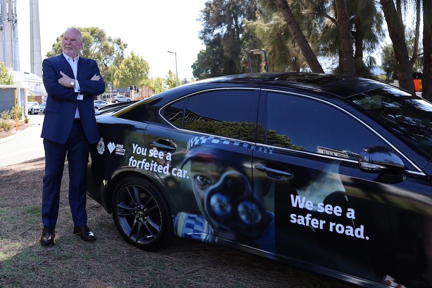 A man in a suit stands with his arms crossed next to a black car with a message about road safety on it. 