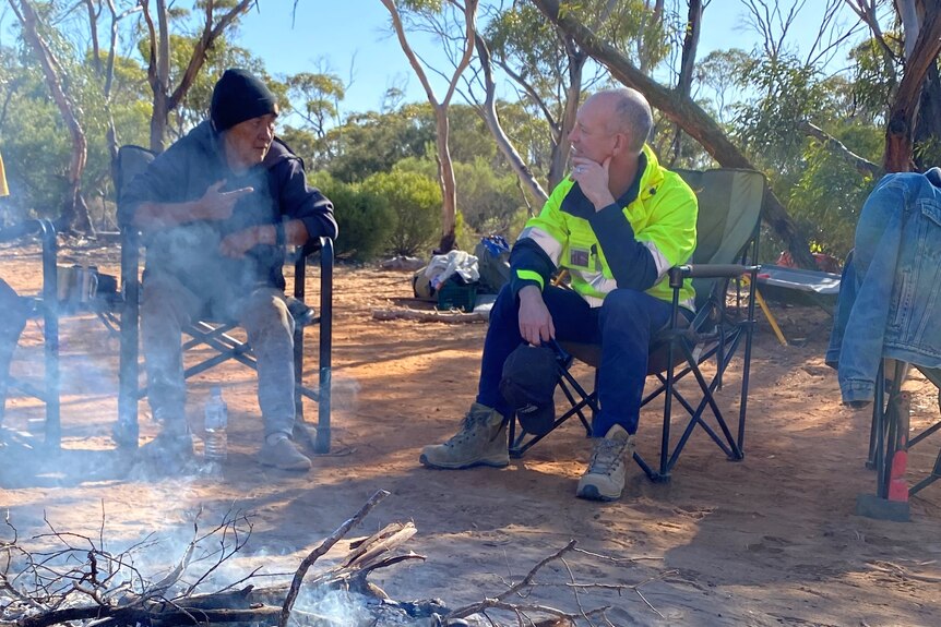 Campfire, kettle in foreground, smoke, woman sitting in a deckchair on left, man in high vis in chair on right