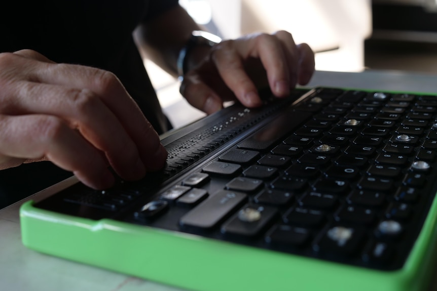 A man's hands feeling the braille on his electronic braille keyboard, which has a green rim.