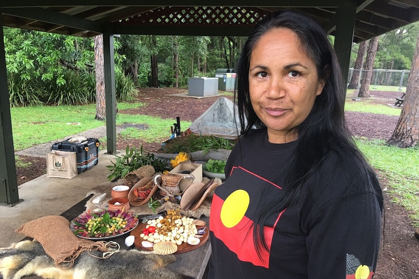 An Indigenous woman wearing an Aboriginal flag T-shirt smiles for the camera in front of a picnic table of fresh foods.