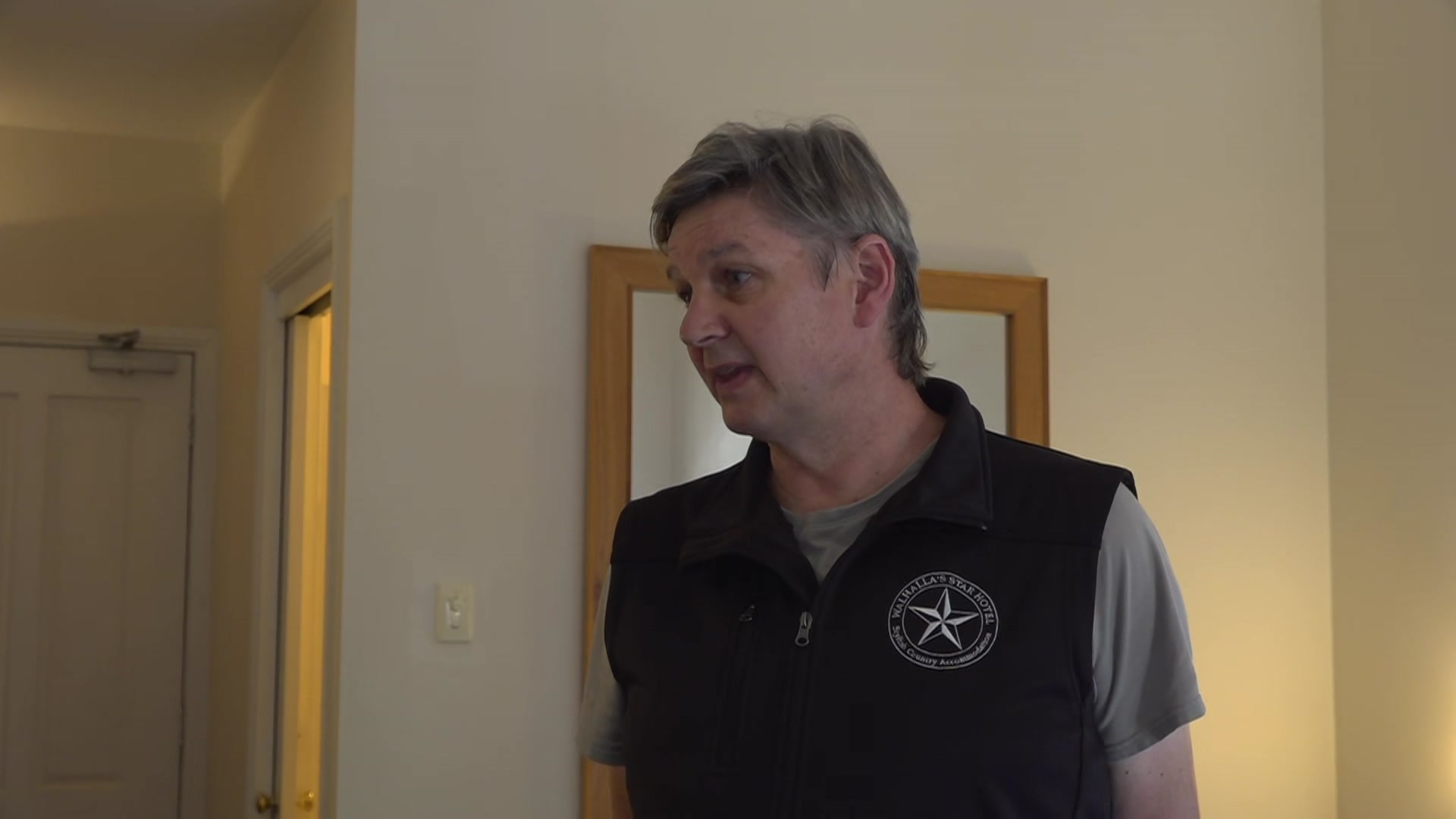 A grey haired man in a black vest stands in a hotel room, talking