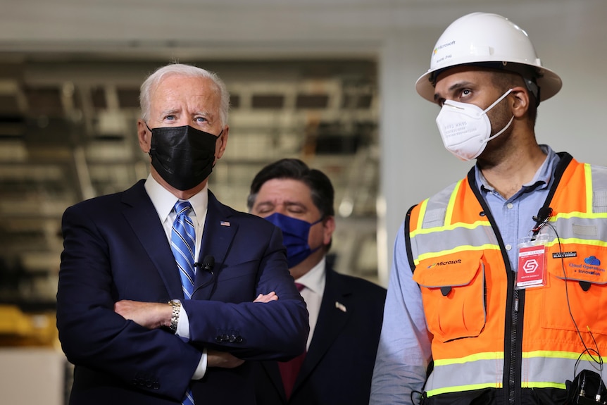 Joe Biden in a mask with his arms crossed next to a man in a high vis vest 