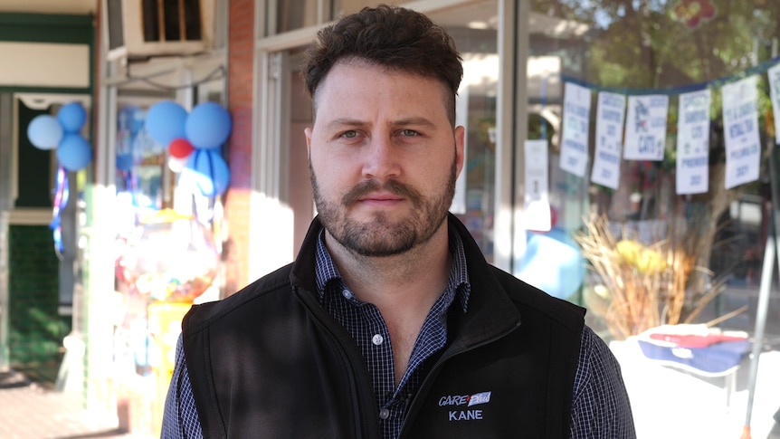 A man with a neat, dark beard and short hair standing outside a pharmacy.