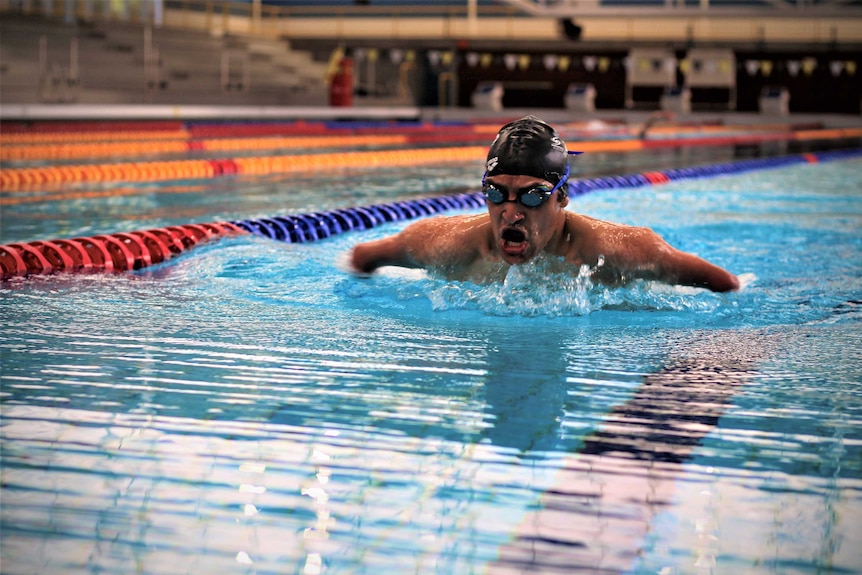 Ahmed Kelly swimming in a pool with goggles and a black swimming cap on. He is mid-stroke.