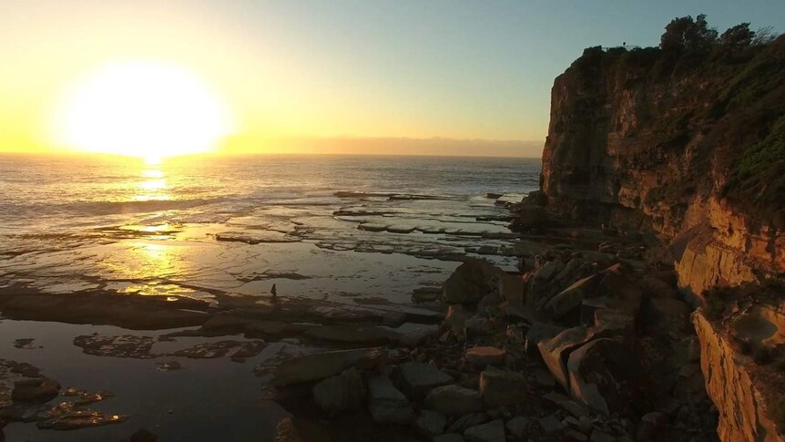 A photo of the Haven at Terrigal. Sun sets over the ocean with rocks and a cliff edge in foreground.