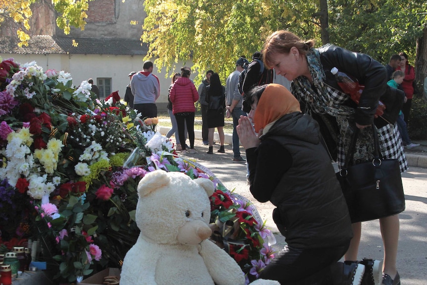 A woman prays next to a memorial adorned with flowers and a large teddy bear, whilst a woman stands next to her.