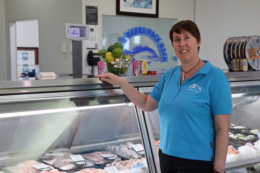 A woman wearing a light blue polo shirt smiles at the camera. She is in the front of a fish fridge, which is full of produce.