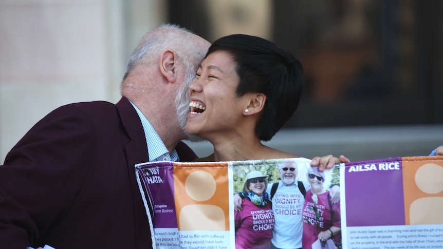 A woman smiles and embraces a man while holding a sign on the steps of parliament