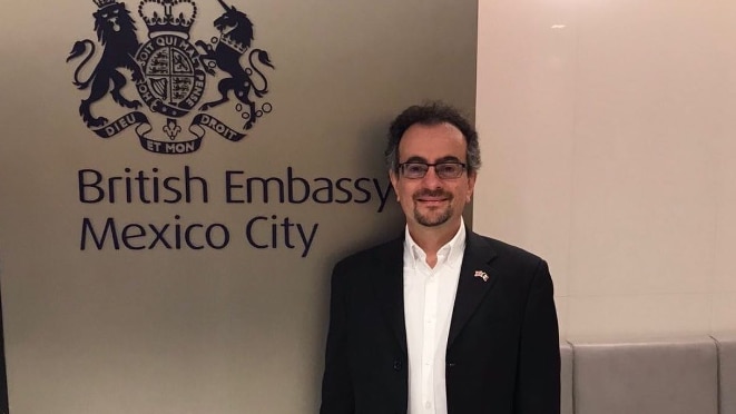 A man with glasses and a goatee stands next to a sign that reads British Embassy Mexico City.