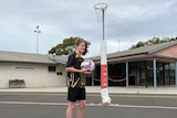 A girl wearing a black T-shirt and shorts holding a netball near a netball ring and clubhouse