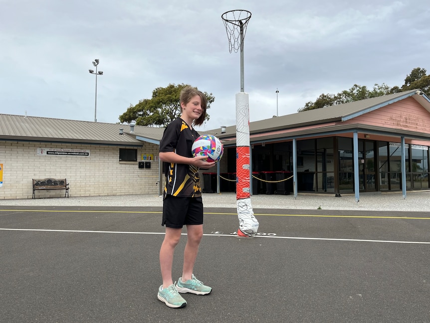 A girl wearing a black T-shirt and shorts holding a netball near a netball ring and clubhouse