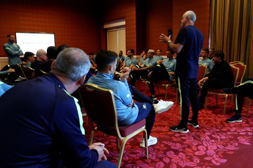 A man wearing navy blue speaks to a meeting room full of soccer players