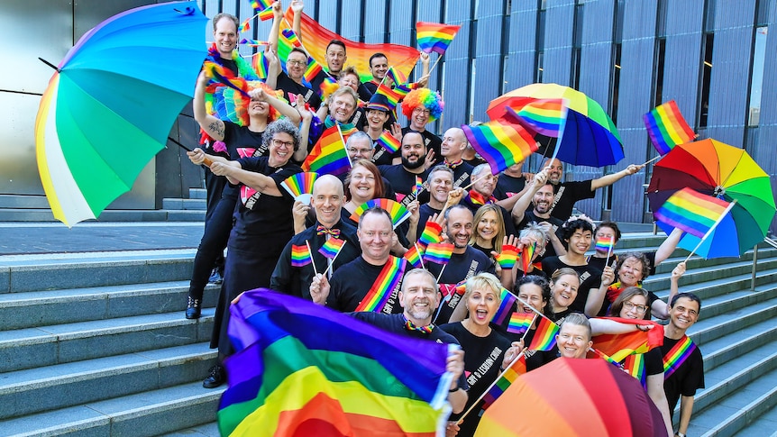 Choristers from the Sydney Gay & Lesbian Choir smiling and holding rainbow flags and umbrellas.