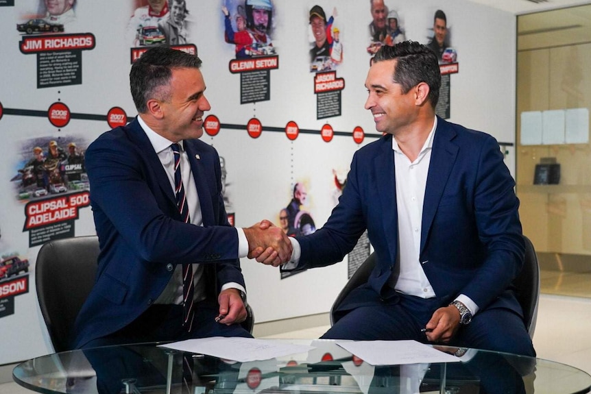 Two men, both wearing suits, shaking hands in front of a wall emblazoned with the Supercars drivers hall of fame.