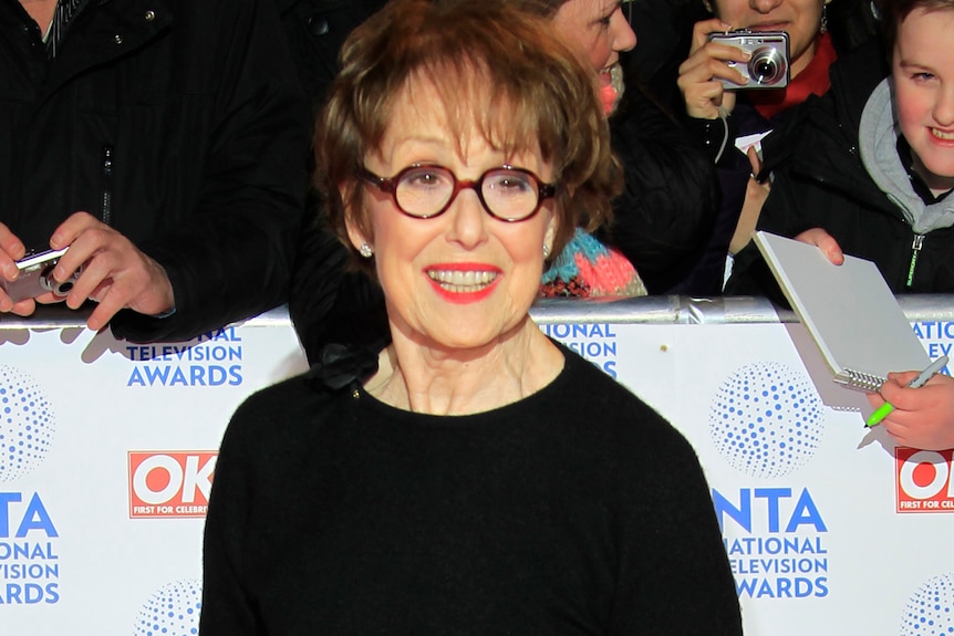 A woman wearing a long black dress with long sleeves and glasses stands on a red carpet. 