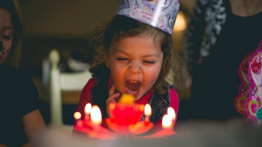 Girl with party hat and cake blowing out candles on a cake for a story about surviving birthday parties