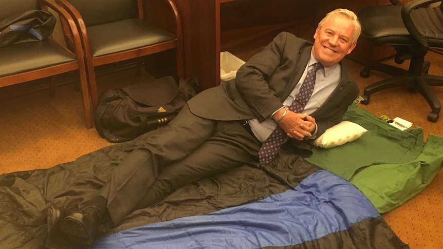 A man in a suit lies down on a swag in a parliamentary office
