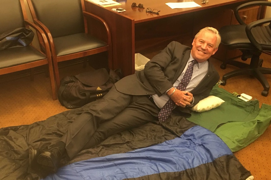 A man in a suit lies down on a swag in a parliamentary office