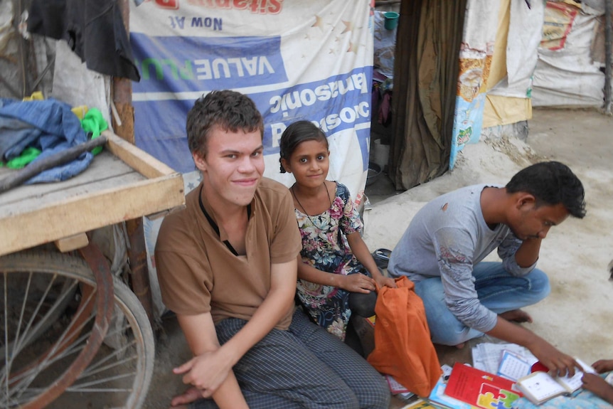 Tom in brown shirt smiles at camera next to young Indian girl. Man in torn light blue shirt reading a book on the right side.