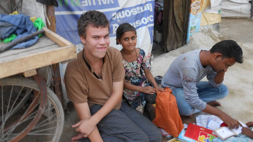 Tom in brown shirt smiles at camera next to young Indian girl. Man in torn light blue shirt reading a book on the right side.