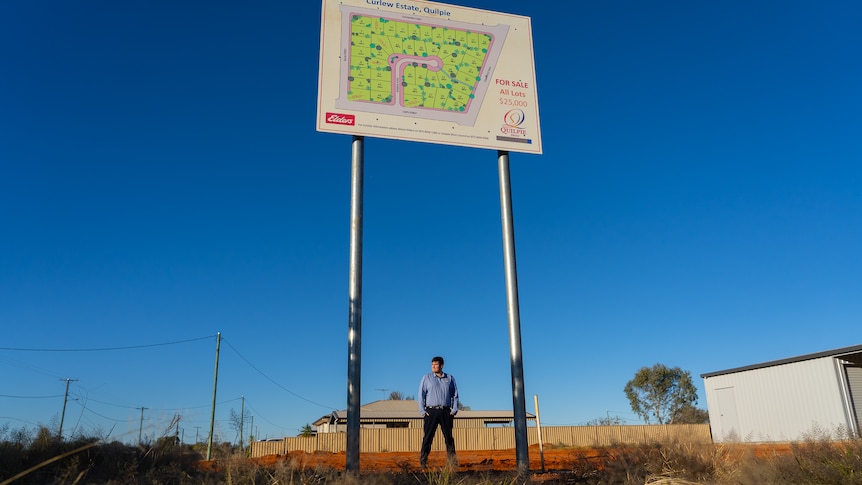 A man stands on a vacant housing block underneath a sign advertising land for sale.