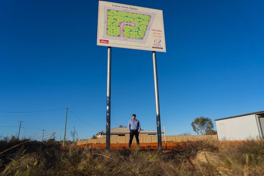 A man stands on a vacant housing block underneath a sign advertising land for sale.