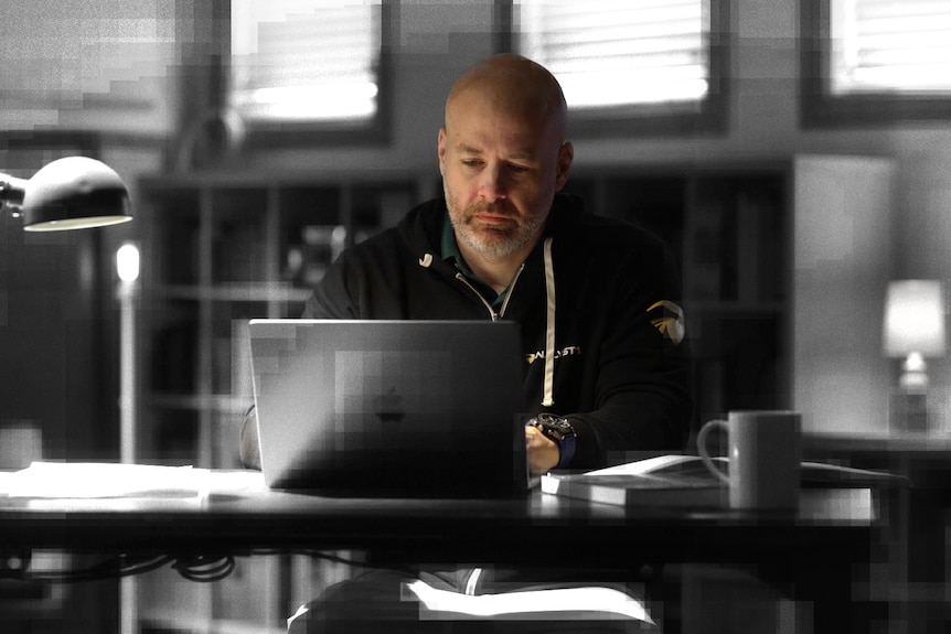A man sits at a desk looking at a laptop in front of him. The picture has been edited to make everything but him black and white