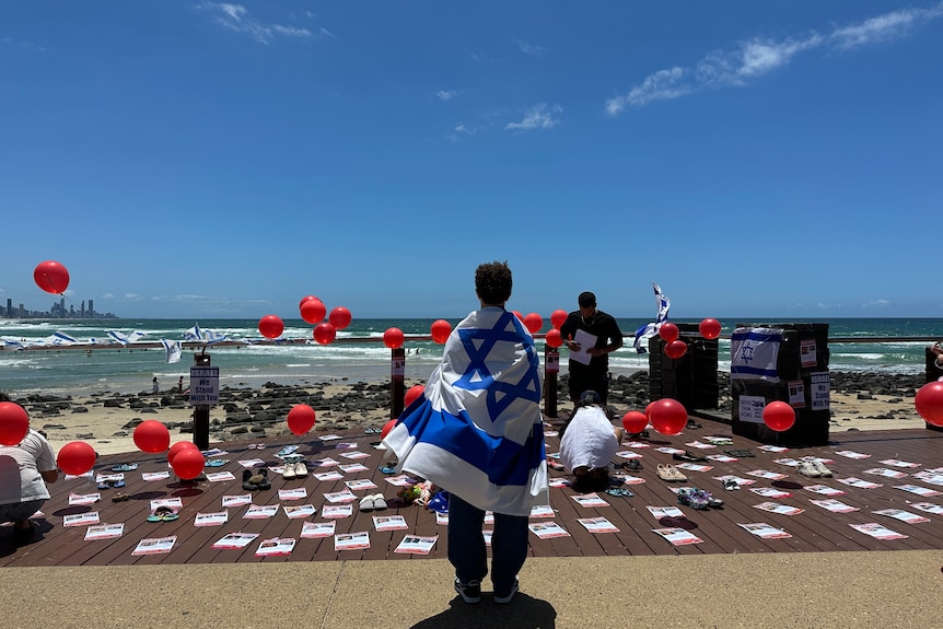 a person wrapped an an israeli flag looks over a pro-israel vigil at the beach at burleigh heads with posters and red balloons