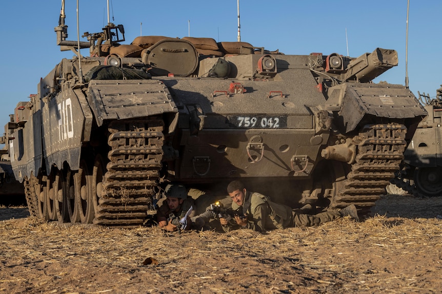 Israeli soldiers take cover under a tank, laying on the ground while holding up their guns.