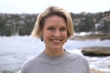 A blonde woman looks at the camera in a coastal setting