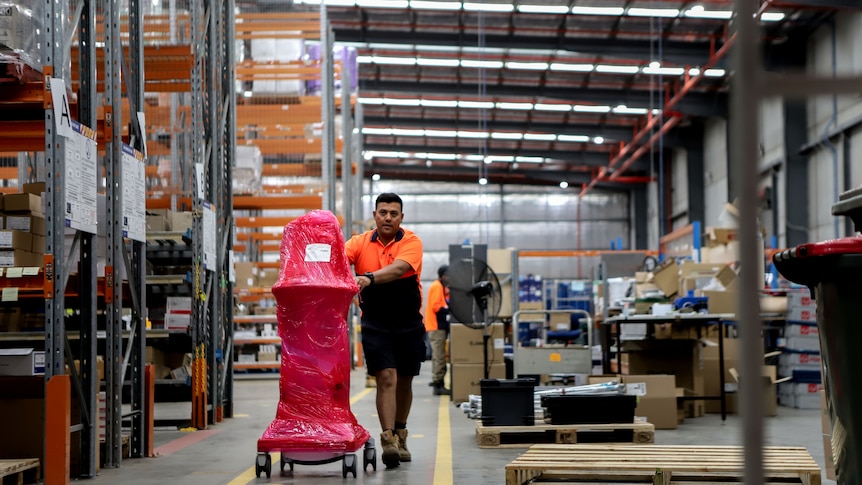 Man in high-vis shirt wheels red plastic wrapped ventilator across warehouse floor next to  large shelves