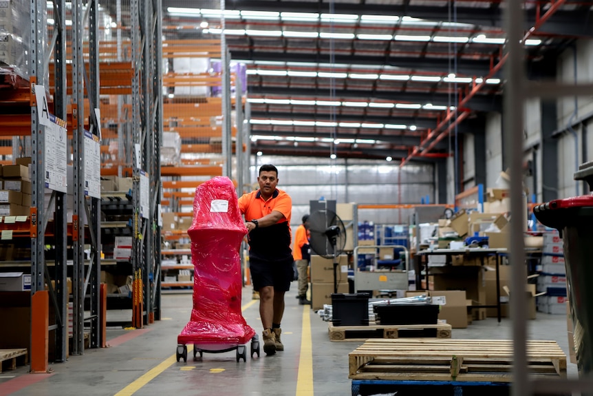 Man in high-vis shirt wheels red plastic wrapped ventilator across warehouse floor next to  large shelves