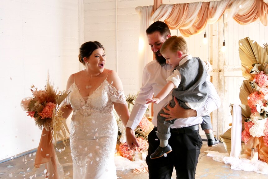 A bride carrying a bouquet and a groom carrying a young boy walk through confetti 