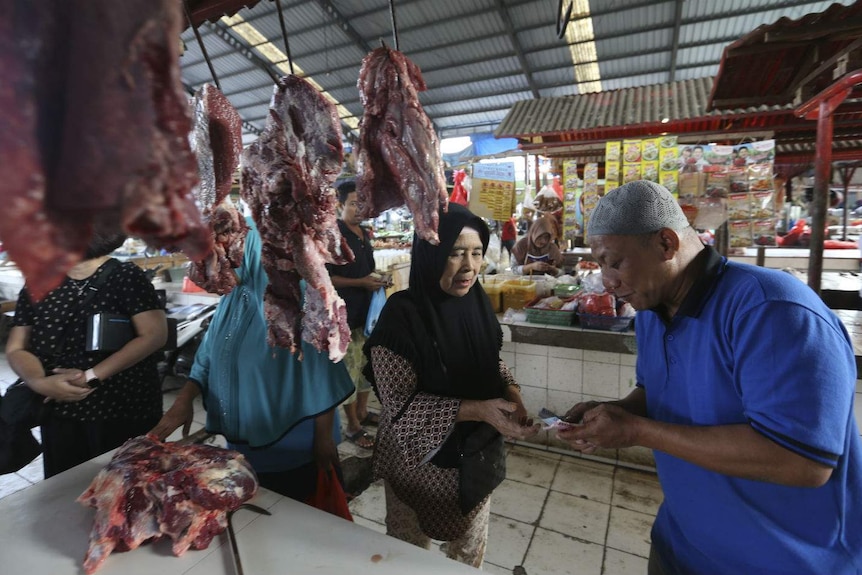 Two women and a male stall holder exchange cash at a market stall lined with meat on hooks at an undercover market.