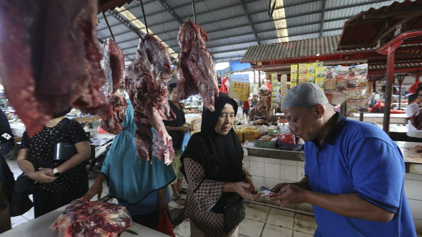 Two women and a male stall holder exchange cash at a market stall lined with meat on hooks at an undercover market.