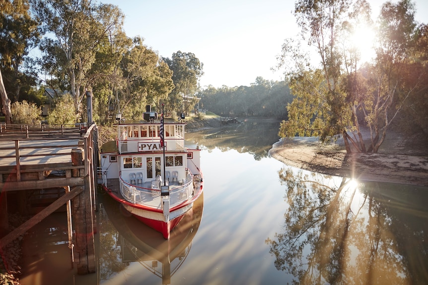 a paddle steamer at a jetty on a wide, tree-lined river.