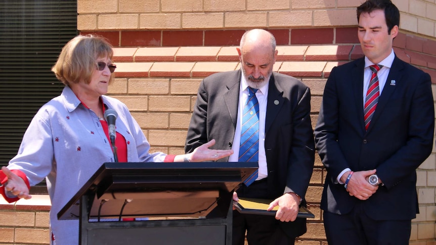 Ann and Graham Brown and John Bale stand at a lectern outside a brick building.