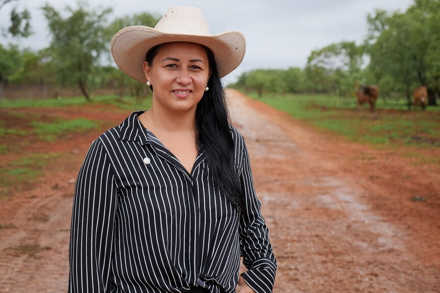 An indigenous woman wearing a black and white pinstripe shirt and white cowboy hat standing on red dirt