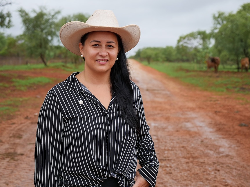 An indigenous woman wearing a black and white pinstripe shirt and white cowboy hat standing on red dirt