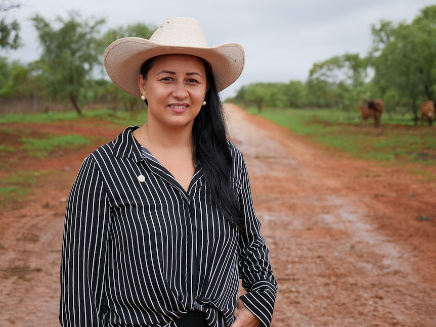 An Indigenous woman wearing a black and white pinstripe shirt and white cowboy hat standing on red dirt