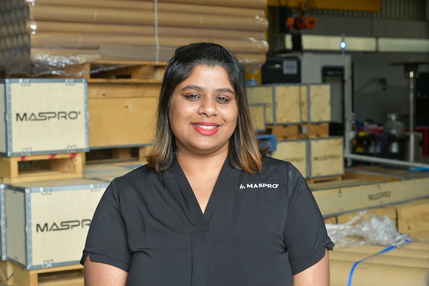 A closeup of a smiling woman in a black blouse who is standing in front of timber boxes.