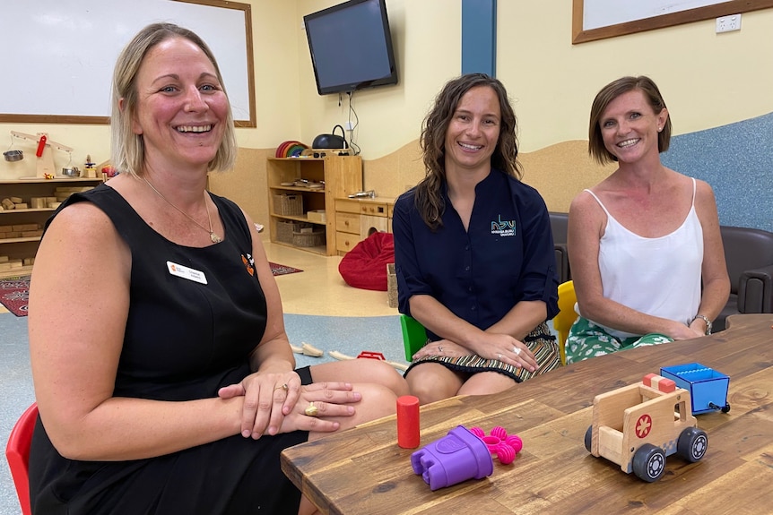Three women smile as they sit on small chairs with toys in front of them