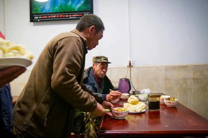 A Chinese man in a cap uses chopsticks to pick up a bun from a plate as a waiter passes by