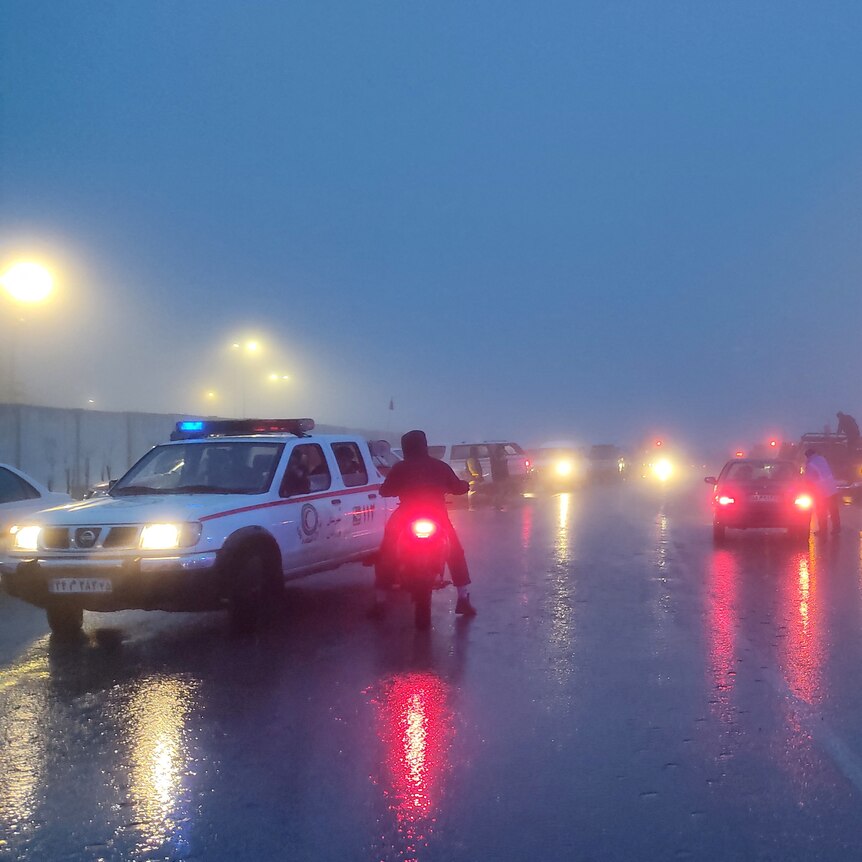 Ambulance and police vehicles are seen in traffic on a foggy, dark stretch of road. 