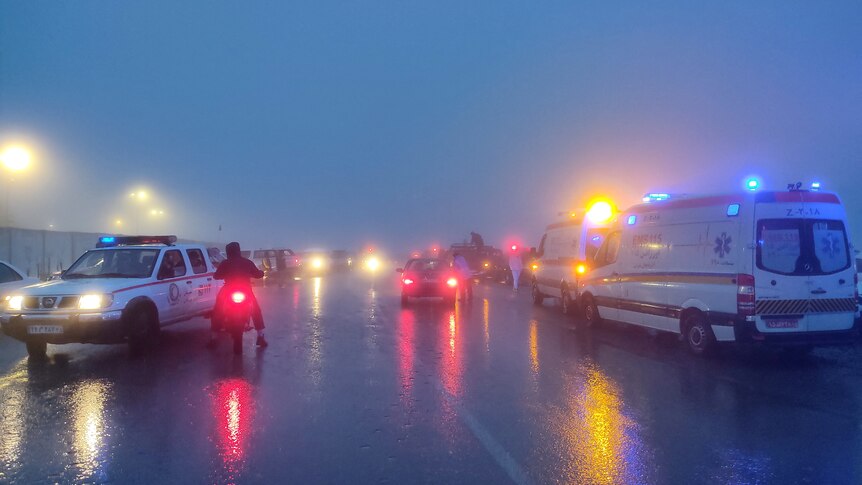 Ambulance and police vehicles are seen in traffic on a foggy, dark stretch of road. 