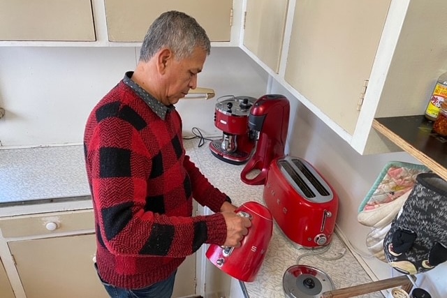 A man in a red and black checkered jumper pours water from a red kettle in a kitchen.