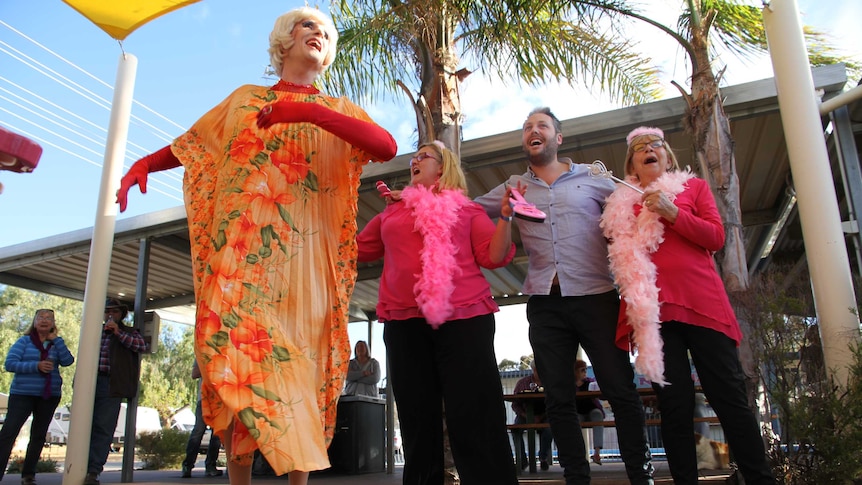 A man dressed in drag sways his arms while two women wearing feather boa and a man dance behind her