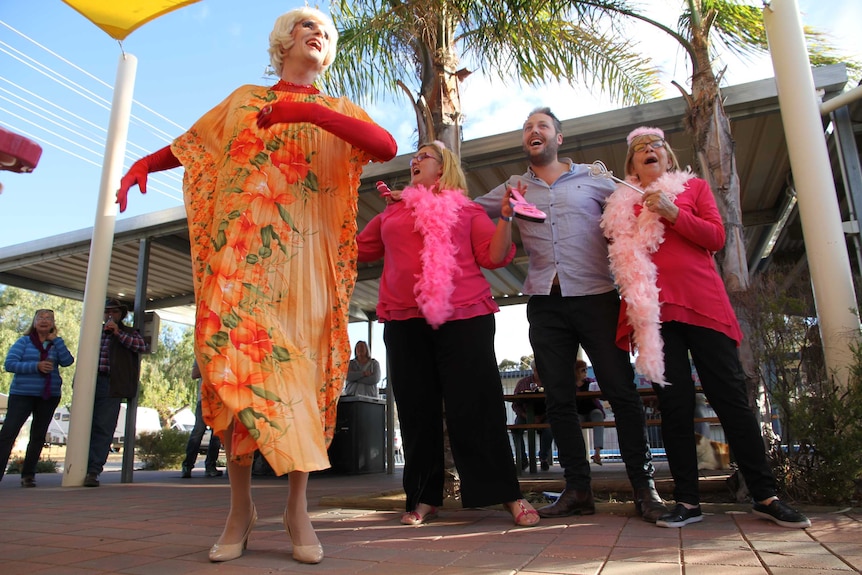 A man dressed in drag sways his arms while two women wearing feather boa and a man dance behind her